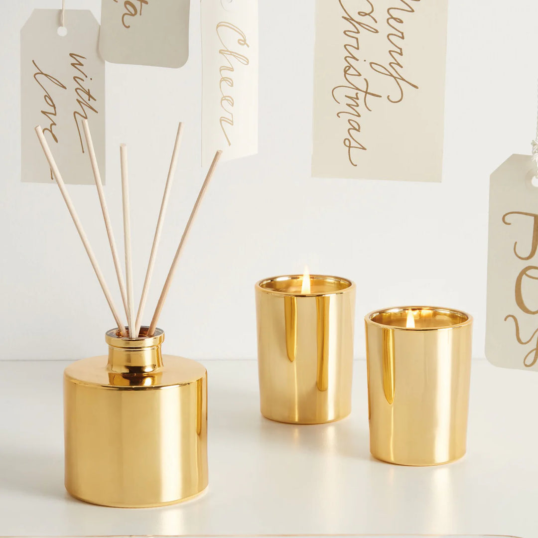 Professional product photograph of a gold home fragrance set on a white tabletop featuring a reflective metallic reed diffuser and two lit votive candles positioned before hanging holiday tags with gold calligraphy