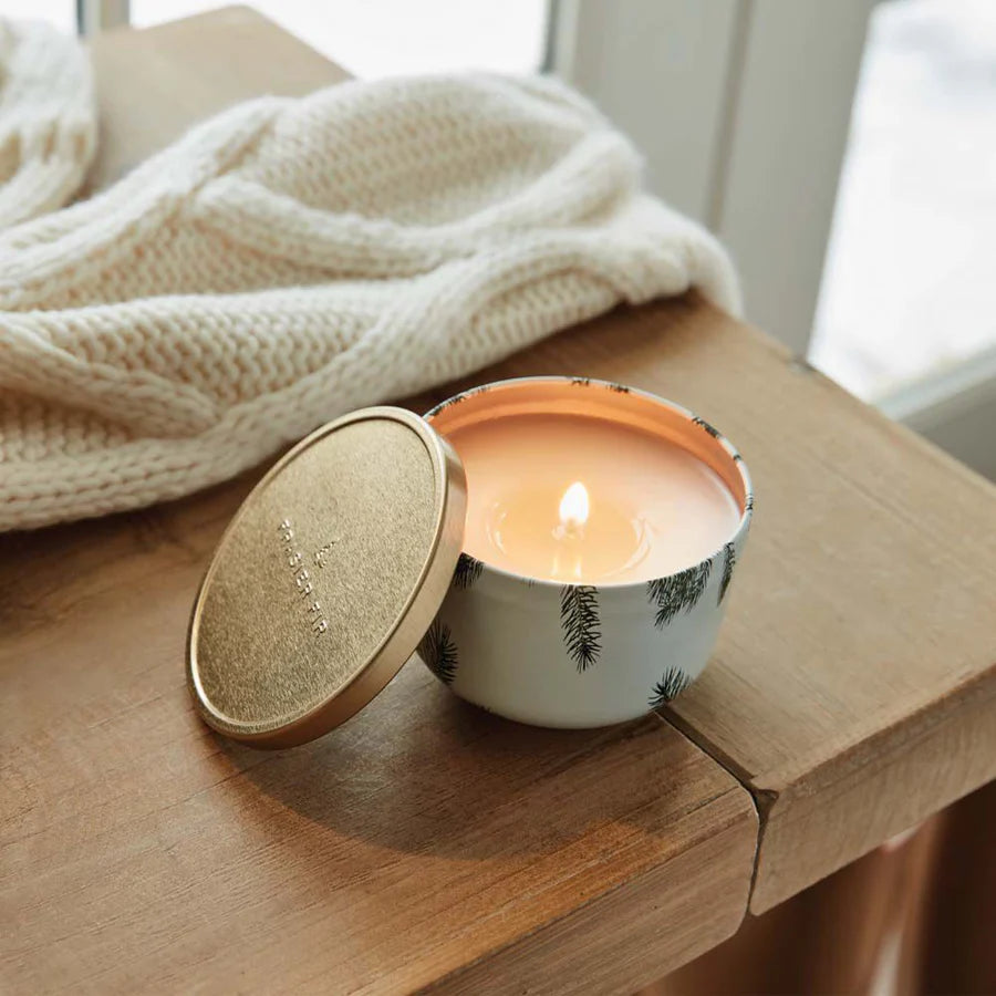 Photograph of a Thymes Frasier Fir scented candle burning in a decorative white tin with pine needle patterns on a wooden table beside its embossed gold lid and a soft white cable-knit sweater