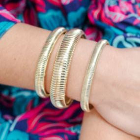 Photograph of three gold-toned ribbed bracelets with a textured snake-like coil design worn on a woman's wrist against a background of vibrant pink and teal floral print clothing