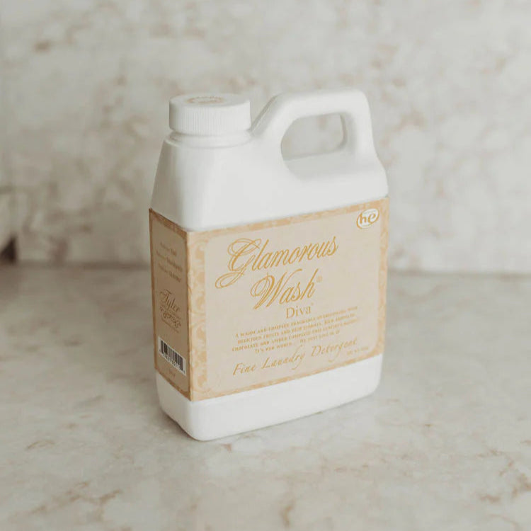 Photograph of a Tyler Candle Co white plastic jug of Diva scent Glamorous Wash laundry detergent on a marble countertop featuring the high-efficiency he logo and elegant gold script on a cream-colored vintage label