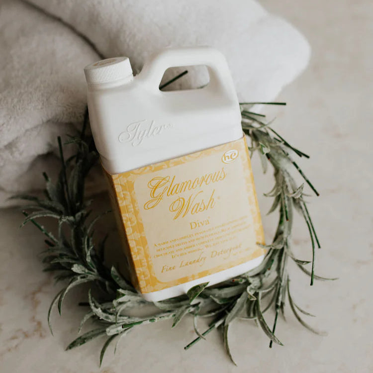 Commercial photograph of a Tyler Candle Company Glamorous Wash Diva laundry detergent bottle nestled in a green rosemary wreath on a marble surface featuring a yellow damask label and soft white towels