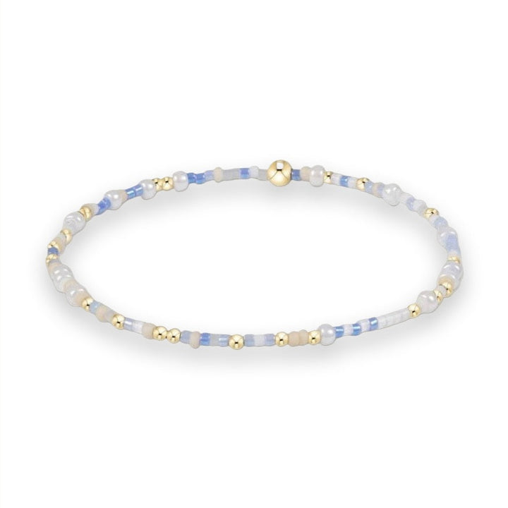 Beaded jewelry bracelet centered on a white background featuring an alternating pattern of periwinkle blue and iridescent white seed beads accented with small polished gold spheres and a single larger gold bead focal point
