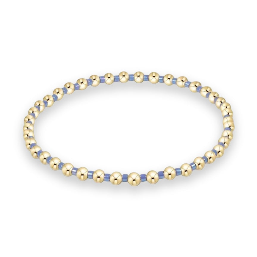 Gold-filled and glass beaded jewelry stretch bracelet centered on a white background featuring polished spherical gold beads and iridescent periwinkle blue cylindrical spacers arranged in a circular design