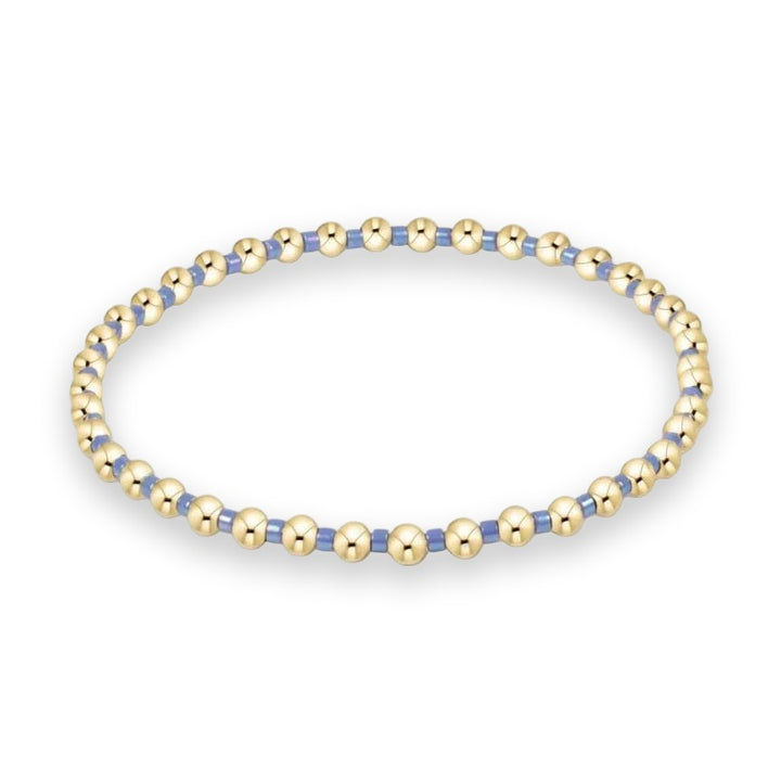 Gold-filled and glass beaded jewelry stretch bracelet centered on a white background featuring polished spherical gold beads and iridescent periwinkle blue cylindrical spacers arranged in a circular design