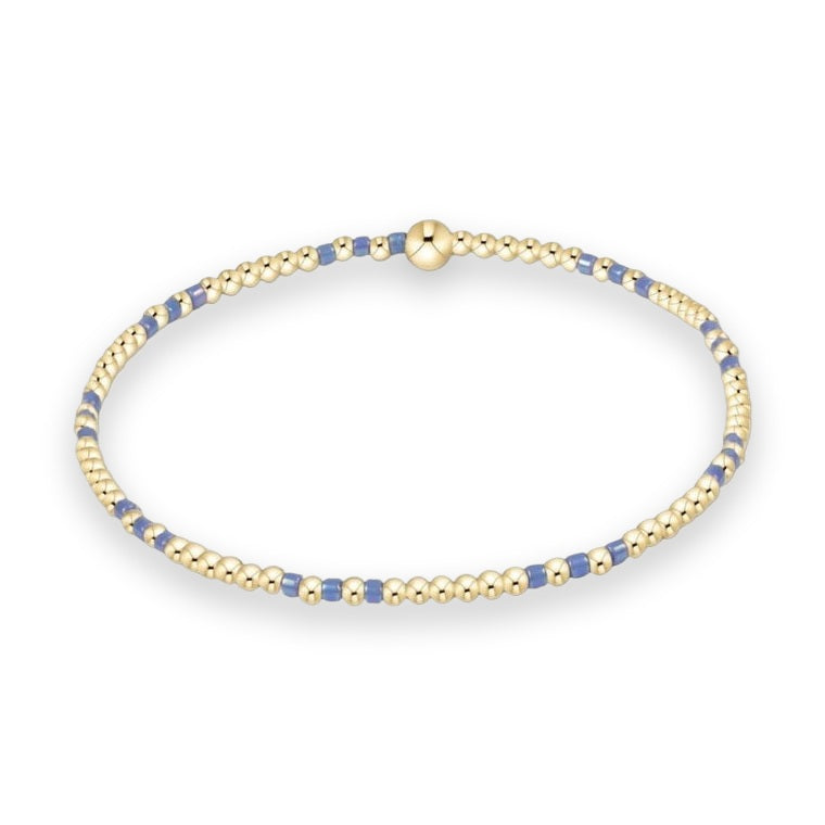 Handcrafted beaded jewelry featuring a delicate gold stretch bracelet on a white studio background designed with alternating polished gold spheres and iridescent periwinkle blue spacers and a single large gold bead focal point
