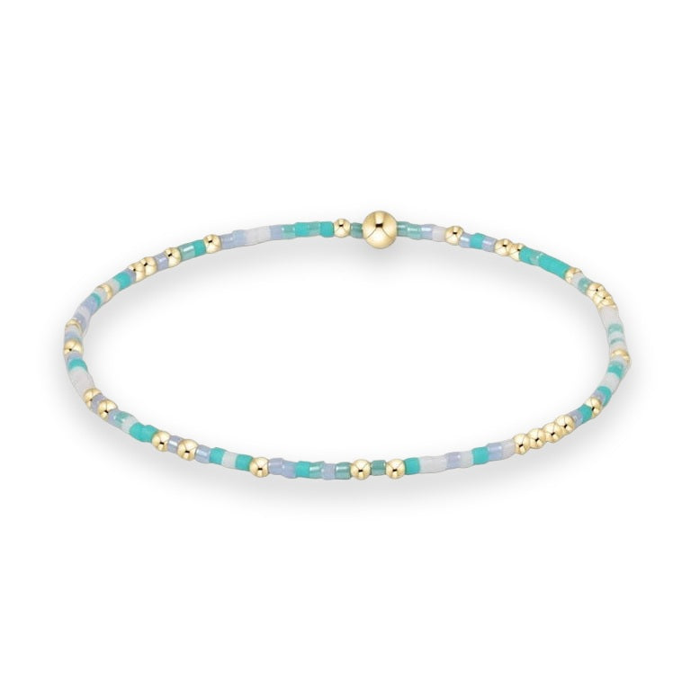 Handcrafted beaded jewelry bracelet centered on a white background featuring a pattern of turquoise, lavender, and white glass seed beads highlighted by polished gold-tone spheres and a central gold ball