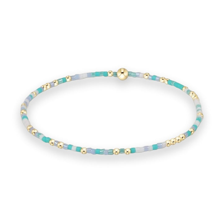 Handcrafted beaded jewelry bracelet centered on a white background featuring a pattern of turquoise, lavender, and white glass seed beads highlighted by polished gold-tone spheres and a central gold ball
