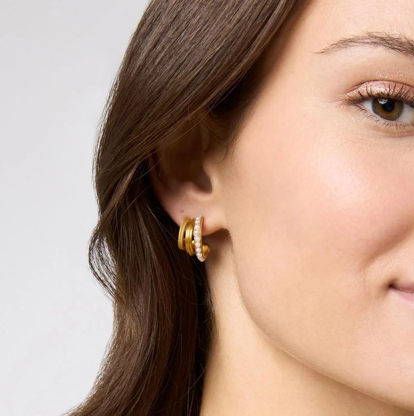 Close-up photograph of a woman wearing a triple-band gold hoop earring in a studio portrait featuring a strand of small white pearls and two textured gold coils complemented by shimmering bronze eye makeup