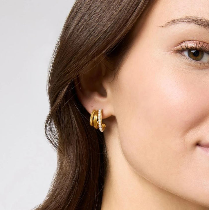 Close-up photograph of a woman wearing a triple-band gold hoop earring in a studio portrait featuring a strand of small white pearls and two textured gold coils complemented by shimmering bronze eye makeup