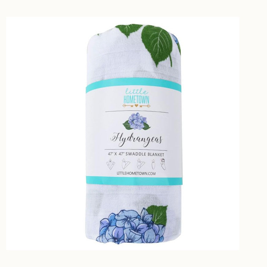 Muslin swaddle blanket by Little Hometown featuring a rolled white fabric with blue hydrangea floral prints and green leaves wrapped in a branded paper label with a teal banner on a white background