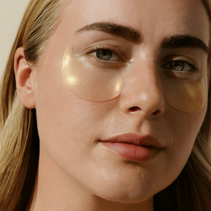 Digital photograph of a woman’s face in a close-up studio setting featuring shimmering gold crescent-shaped under-eye patches, defined dark eyebrows, and a soft neutral lip color