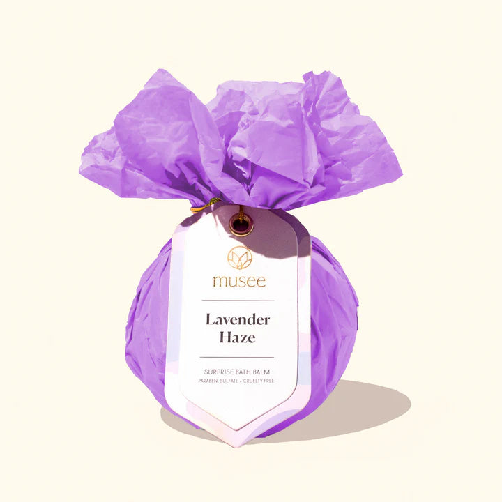 Photograph by Musee of a Lavender Haze surprise bath balm wrapped in ruffled purple tissue paper with a white arched tag featuring gold lettering and a long soft shadow on a neutral cream background