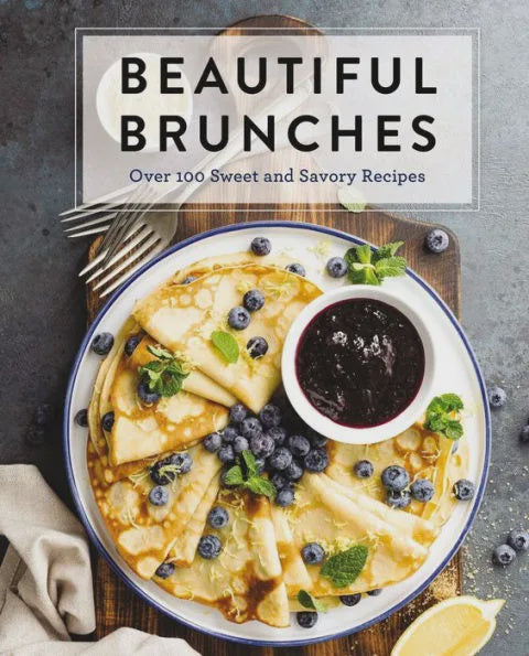 Food photography of a cookbook cover titled Beautiful Brunches featuring golden folded crepes on a blue-rimmed plate garnished with fresh blueberries, vibrant mint leaves, and yellow lemon zest on a rustic wooden board