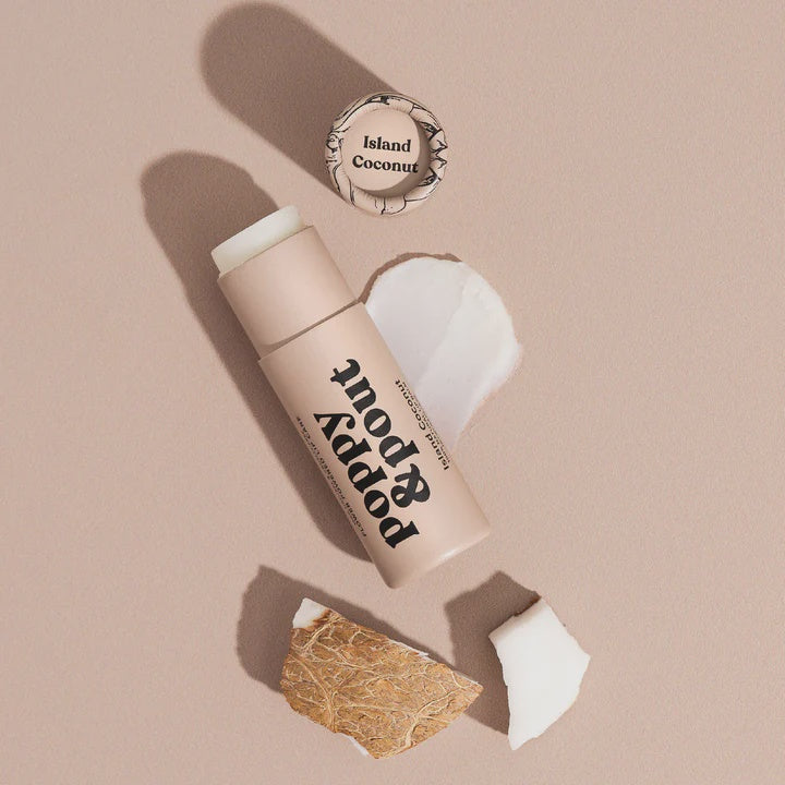 Product photograph by Poppy & Pout of an Island Coconut lip balm on a neutral beige surface featuring the brand logo, a creamy white balm smear, and chunks of textured brown coconut husk