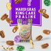 Digital graphic advertisement by New Orleans Famous Praline Company featuring a Mardi Gras King Cake Praline package surrounded by floating pecans, festive purple, green, and gold doubloons, and a swirling gold ribbon against a vibrant purple backdrop
