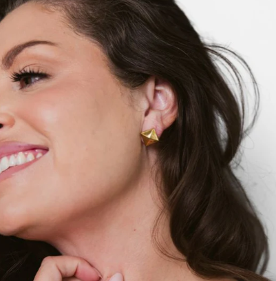 Digital photograph of a smiling woman with dark wavy hair in a studio setting highlighting a gold pyramid stud earring, soft pink lipstick, and a radiant smile