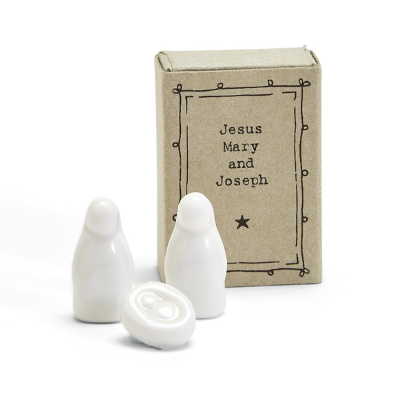 Porcelain miniature nativity set by East of India featuring smooth white glazed figurines of Mary, Joseph, and baby Jesus displayed on a white background next to a rustic kraft matchbox with a hand-drawn border and a single black star