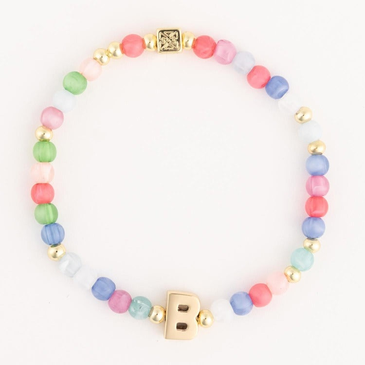 Beaded jewelry bracelet showcases multi-colored pastel cat-eye beads and gold-tone spacers featuring a large gold-tone capital letter B charm and an ornate square bead on a plain white background