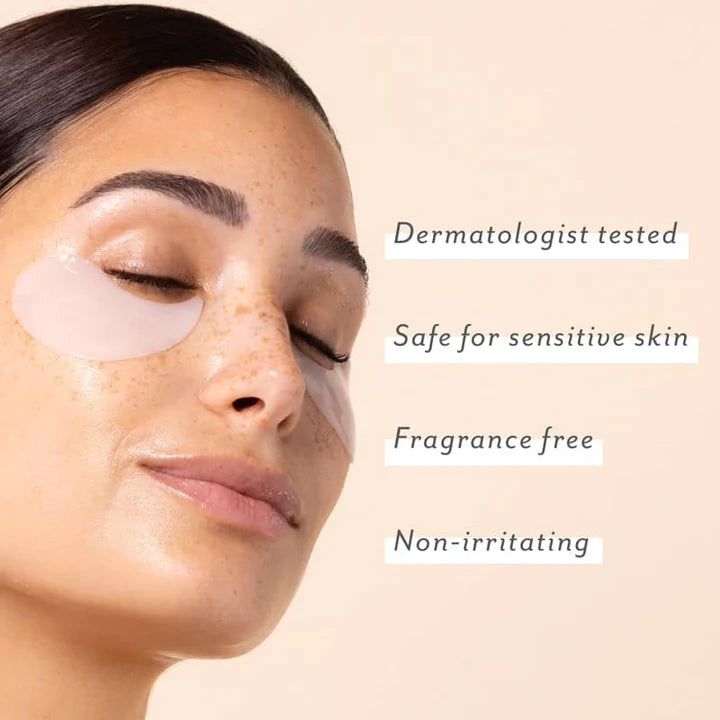 Close-up studio photograph of a woman with freckled skin and a glowing complexion wearing translucent white under-eye patches beside four white text labels for dermatologist tested and fragrance free against a soft peach background