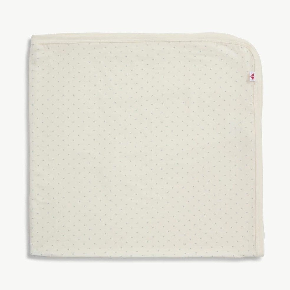 Photograph of a MORI cream-colored textile baby blanket on a white background featuring a tiny grey polka dot pattern, a reinforced hemmed border, and a small red logo tag on the side edge
