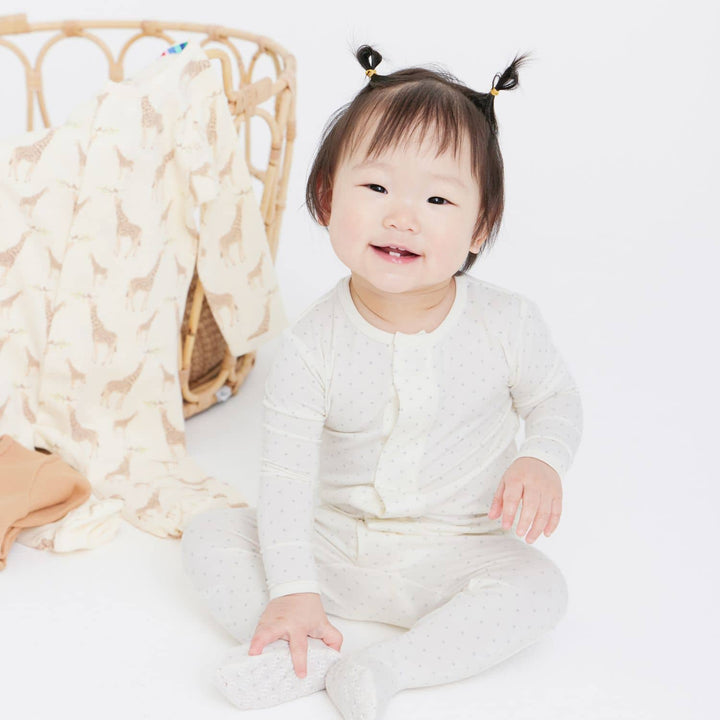 Digital photograph of a smiling East Asian baby girl in a bright white studio featuring two tiny top-knot pigtails, cream-colored dotted pajamas, and a giraffe-patterned blanket draped over a woven rattan basket