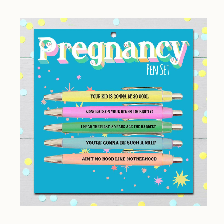 Product photograph of a Fun Club Pregnancy Pen Set on a light wood surface featuring five pastel-colored pens with humorous slogans including Congrats on your recent sobriety and Ain't no hood like motherhood against a teal card with retro 3D typography and scattered confetti