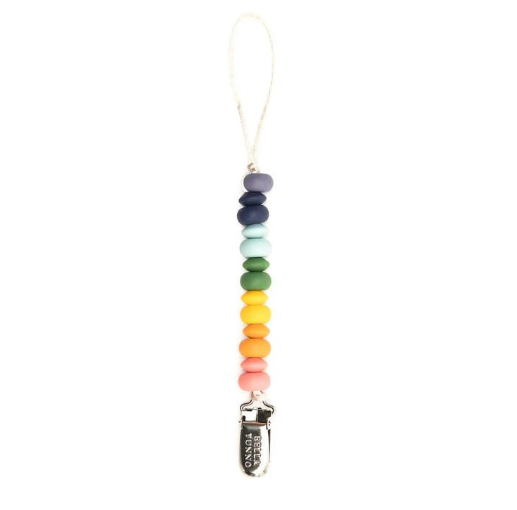 Silicone and metal pacifier clip by Bella Tunno featuring a vertical strand of matte rainbow-colored beads ending in a silver metal clip engraved with the brand name against a plain white background