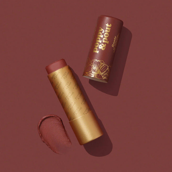 Commercial studio photograph for Poppy &amp; Pout of a Roxie tinted lip balm tube on a dark red background featuring gold foil lettering, an intricate floral design, and a creamy product swatch