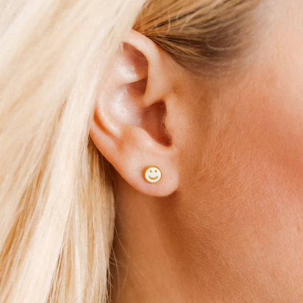 Digital photograph of a small gold and white enamel smiley face stud earring worn on a human earlobe featuring a minimalist design and strands of light blonde hair