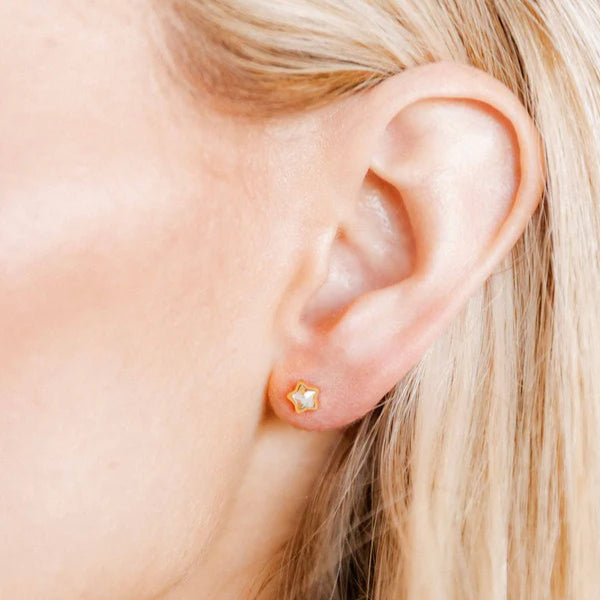 Jewelry photograph of a dainty gold-bordered star stud earring featuring a white pearlescent inlay worn on a human earlobe and framed by light blonde hair