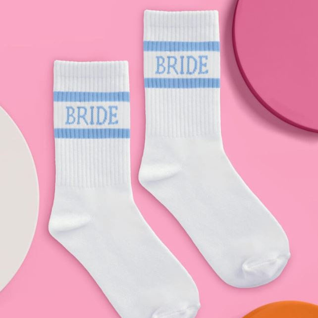 Commercial product photograph of white crew socks with light blue horizontal stripes and the word BRIDE in serif font laid flat on a bright pink background featuring ribbed textures and colorful circular accents