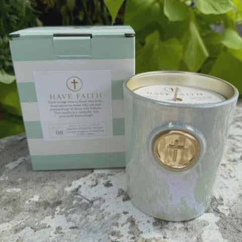 Ceramic Have Faith scented candle and its mint-green striped box on a stone surface outdoors, featuring an iridescent light blue jar with a gold cross medallion and a matching gold-trimmed rim around the white wax interior