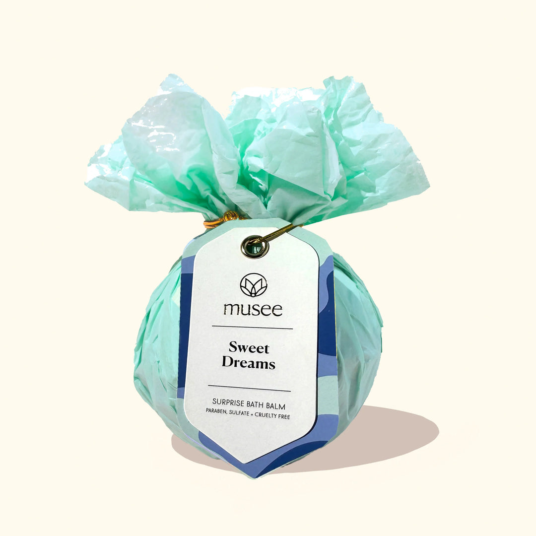 Photograph of a Musee Sweet Dreams Surprise Bath Balm wrapped in ruffled mint green tissue paper and secured with a gold wire tie and a white tag with blue waves on a neutral cream background