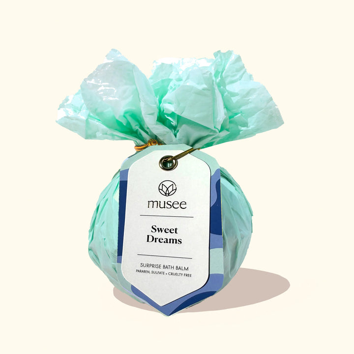 Photograph of a Musee Sweet Dreams Surprise Bath Balm wrapped in ruffled mint green tissue paper and secured with a gold wire tie and a white tag with blue waves on a neutral cream background