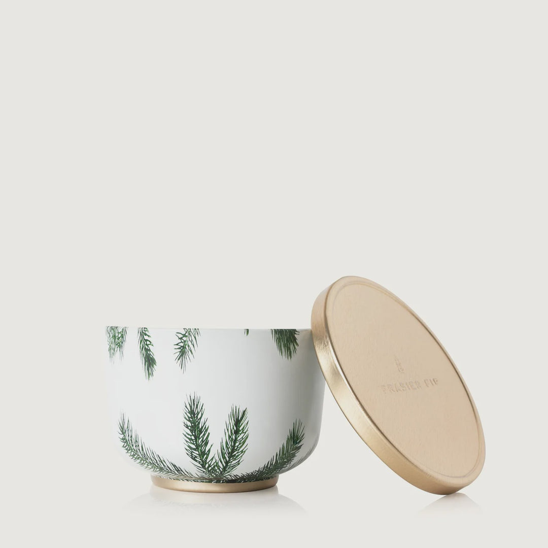 Ceramic Thymes Frasier Fir candle jar on a white background featuring green pine branch illustrations, a metallic gold base, and an embossed gold lid leaning against the container