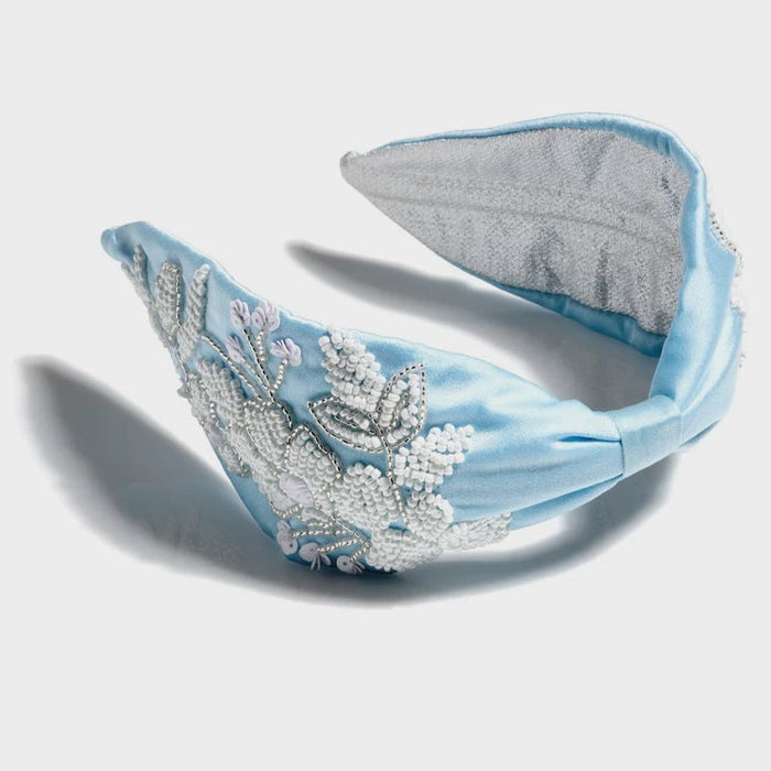 Hand-embroidered satin light blue knotted headband featuring intricate white seed bead floral clusters, shimmering silver metallic thread, and a textured grey lining on a neutral white background