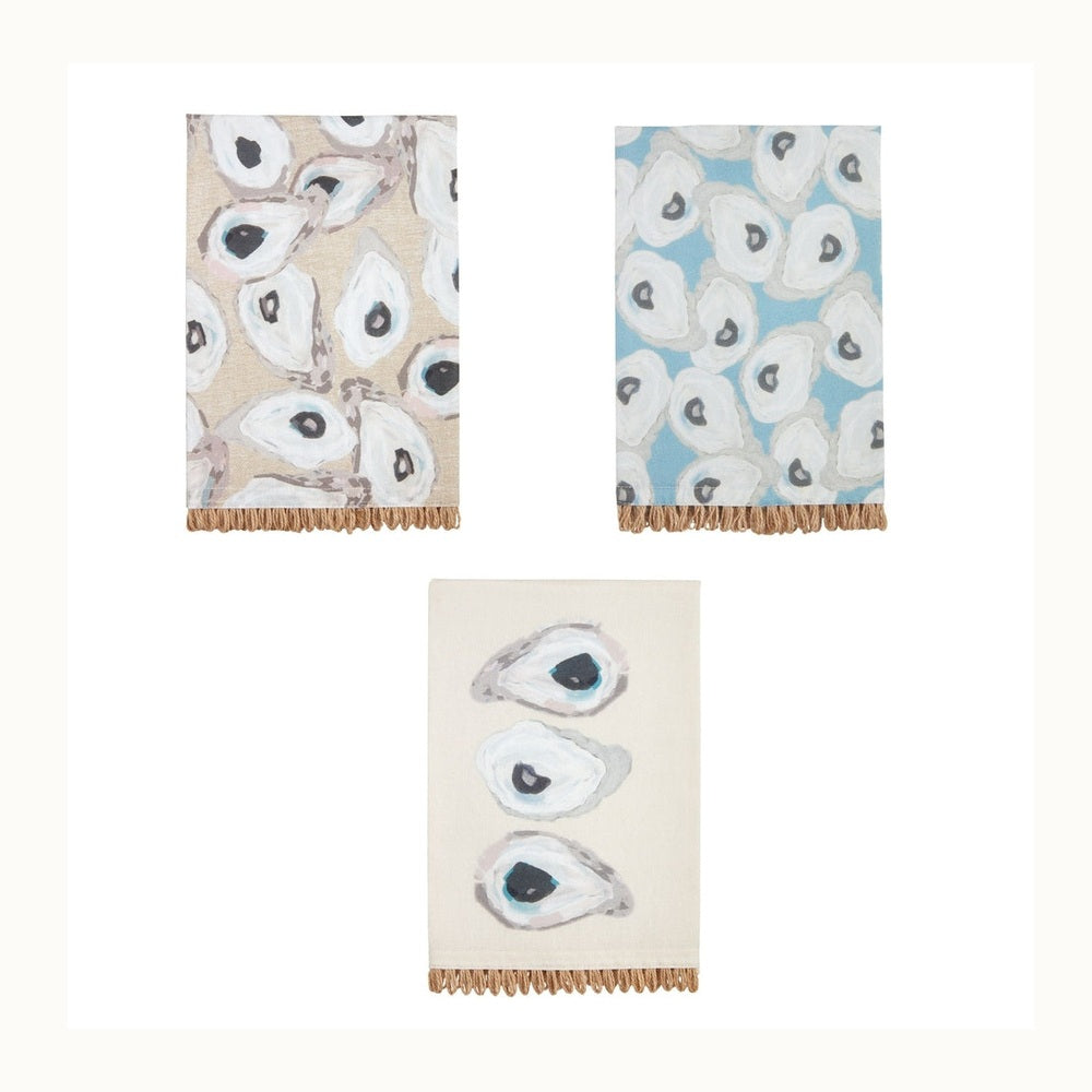 Painted cotton hand towels by Mud Pie featuring coastal oyster shell patterns in soft blue and tan palettes with textured braided jute fringe along the bottom edges