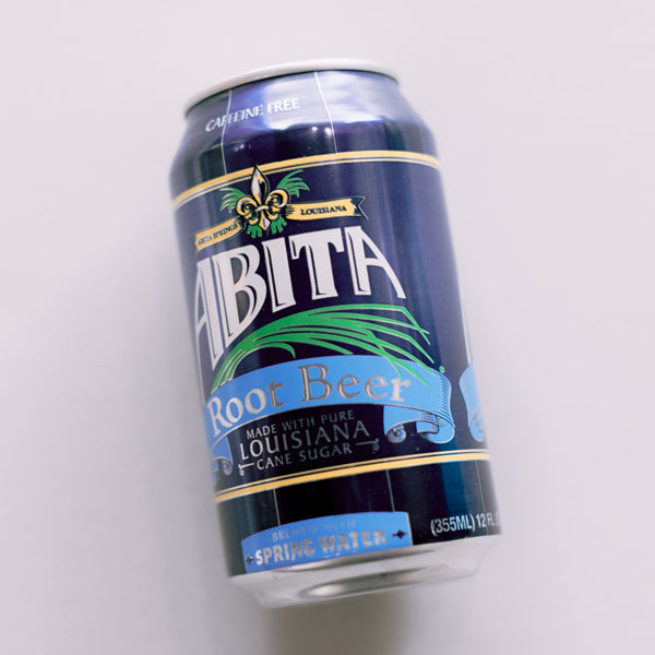 Photograph of an Abita Root Beer can from Louisiana featuring the iconic gold fleur-de-lis symbol, light blue ribbon banners, and navy blue vertical stripes against a white background