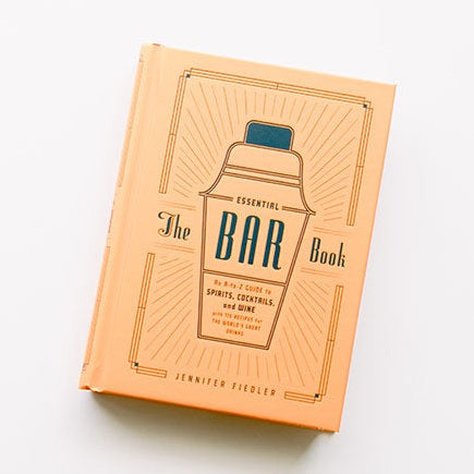 Hardcover book by Jennifer Fiedler titled The Essential Bar Book on a white surface featuring a peach-orange cover with a navy blue Art Deco cocktail shaker illustration and radiating geometric sunburst lines