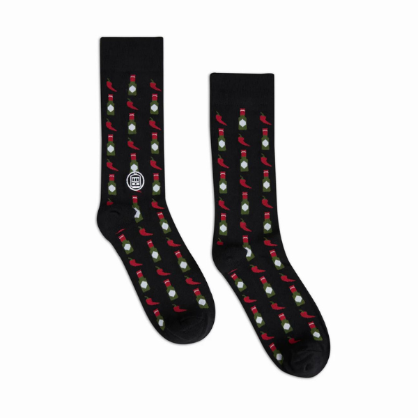 Graphic crew socks by NOLA Couture featuring a black fabric base decorated with a repeating pattern of vibrant red chili peppers, green-capped hot sauce bottles, and the iconic white St. Charles Streetcar emblem from New Orleans