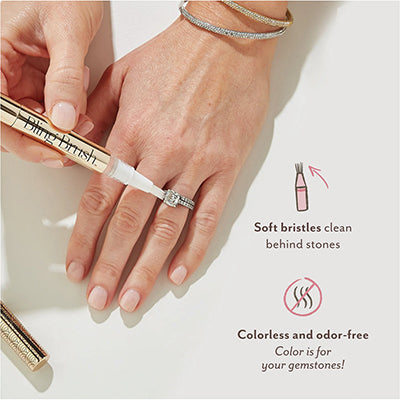 Commercial product photograph of a gold-toned Bling Brush pen cleaning an emerald-cut diamond ring on a hand featuring a light pink manicure and stacked silver bangles