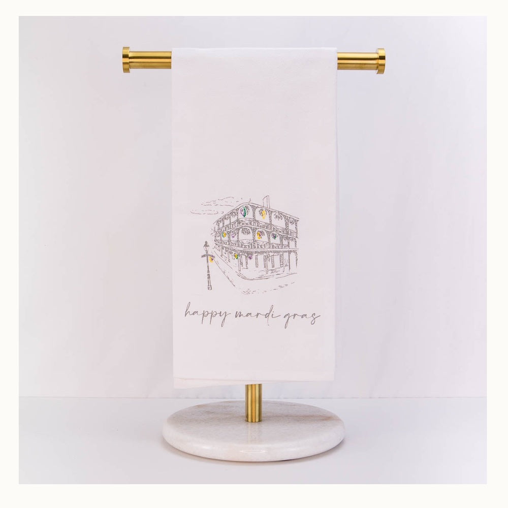 Illustrated white tea towel featuring a sketch of a New Orleans French Quarter building with iconic wrought-iron balconies decorated with colorful purple, green, and gold beads and cursive script reading happy mardi gras