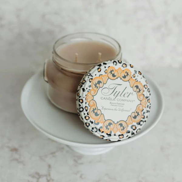 Commercial product photograph by Tyler Candle Company of a double-wick jar candle on a white ceramic pedestal against a marble background featuring a leopard print lid with gold scrollwork and brand typography