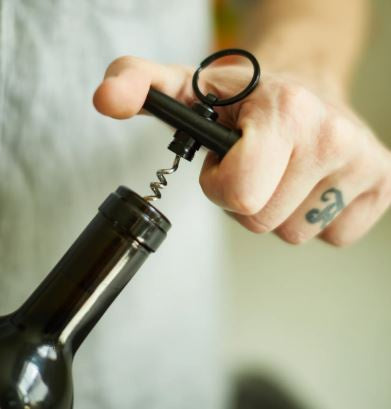Photograph of a hand with a script finger tattoo using a black T-shaped manual corkscrew to open a dark glass wine bottle indoors featuring a circular metal ring and a coiled silver screw