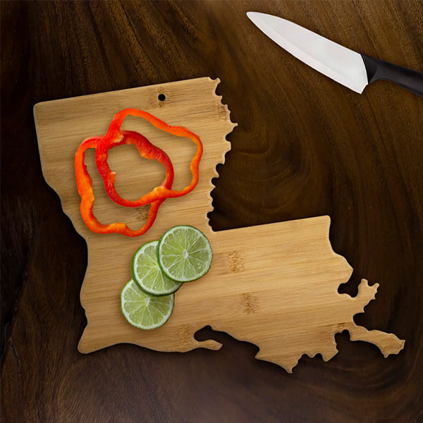 Bamboo cutting board shaped like the state of Louisiana on a dark wood background featuring vibrant red bell pepper rings, fresh lime slices, and a white ceramic knife