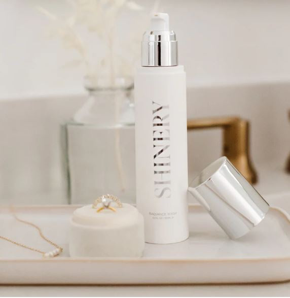 Photograph of Shinery Radiance Wash jewelry cleaner on a bathroom vanity tray featuring a white pump bottle with reflective silver lettering, a gold diamond ring on a velvet stand, and a delicate gold necklace