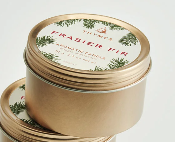 Photograph of Thymes Frasier Fir aromatic candles in brushed gold tins on a white background featuring green fir needle illustrations and the signature pine tree logo alongside red serif lettering
