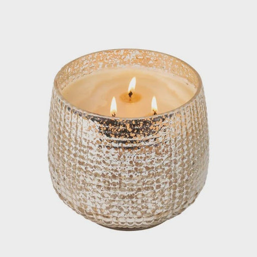 Studio photograph of a three-wick scented candle in a textured champagne mercury glass jar on a neutral background featuring three flickering orange flames and a shimmering metallic hobnail finish