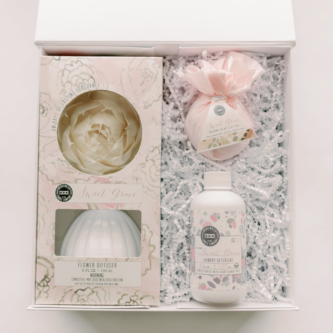 Photograph of a Bridgewater Candle Company Sweet Grace gift set featuring a white ceramic flower diffuser, a pink tissue-wrapped bath bomb, and a laundry detergent bottle nestled in white crinkle paper fill within a gift box