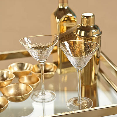 Digital photograph of an elegant gold barware set on a mirrored tray featuring two hammered glass martini glasses with gold rims and a matching polished gold cocktail shaker